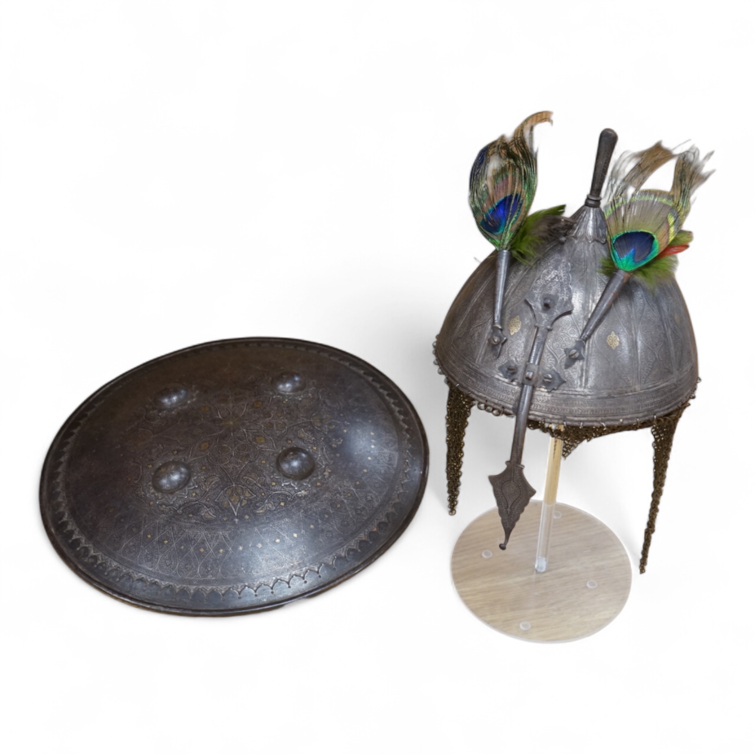 A mid to late 19th century Indian helmet and matching shield, covered with silver koftgari decoration, the helmet with sliding nasal bar and twin plume sockets surmounted by a third plume socket, the skull fluted like a
